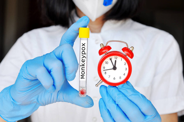  A medical worker wearing surgical face mask and gloves holding test tube, diagnosis and monkeypox research.Holding red alarm clock, It's five to twelve, time is running out - Photo, Image