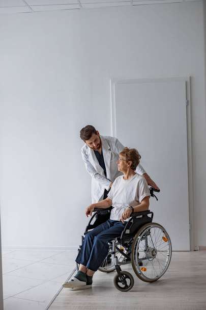 診療所のホールで車椅子で患者を運ぶプロの男性医師。高品質の写真 - 写真・画像