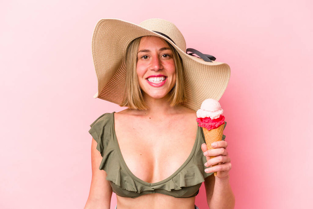 Young caucasian woman going to the beach eating ice cream isolated on pink background - Photo, Image