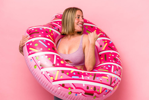 Mujer caucásica joven sosteniendo un colchón de aire aislado en puntos de fondo rosa con el dedo pulgar lejos, riendo y sin preocupaciones. - Foto, imagen