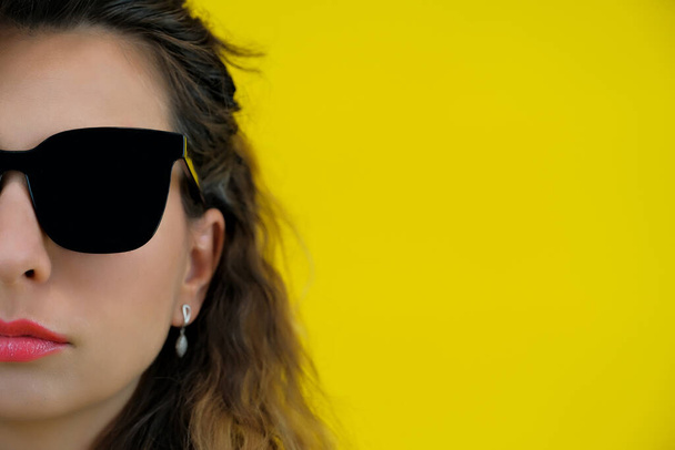 Close-up portret van een mooi meisje op een gele achtergrond. Een vrouw met een zwarte bril ziet er attent uit. menselijke emoties - Foto, afbeelding