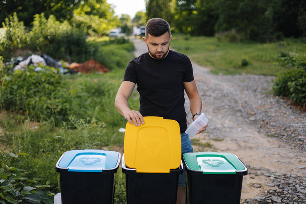 Hombre tirando bolsa de plástico en la papelera de reciclaje en el patio trasero. Diferentes colores en contenedores de plástico. Verde, azul y amarillo. - Foto, imagen