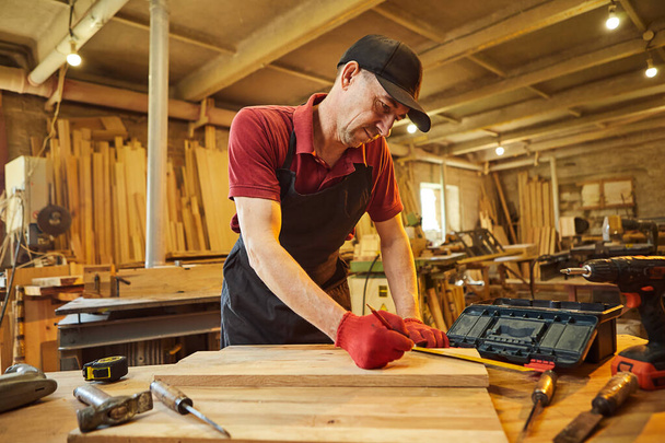 Ξυλουργός που εργάζεται με ένα ξύλο, σήμανση σανίδα με ένα μολύβι και τη λήψη μετρήσεων για να κόψει ένα κομμάτι ξύλου για να κάνει ένα κομμάτι των επίπλων σε ένα εργαστήριο ξυλουργικής - Φωτογραφία, εικόνα