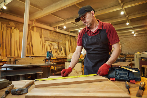 Carpintero trabajando con una madera, marcando tablón con un lápiz y tomando medidas para cortar un trozo de madera para hacer un mueble en un taller de carpintería - Foto, Imagen