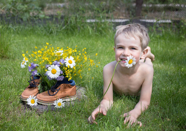 Ein kleiner Junge liegt im Gras, neben einem gesammelten Strauß Wildblumen, der in Stiefeln steht. Freude, positive Atmosphäre, glückliche Kindheit. Hallo Sommer. Überraschungsgeschenk für Mama. Gehen. Tag der Erde - Foto, Bild