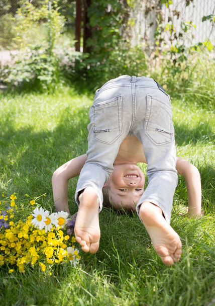bosak bawi się na dworze i radośnie stoi do góry nogami na trawie. Aktywne gry dla dzieci w lecie, zdrowy tryb życia. Przedszkolak się bawi, cieszy życiem. Skupienie selektywne - Zdjęcie, obraz