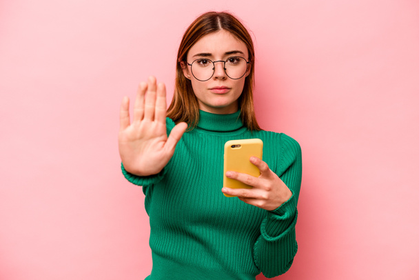 Jonge blanke vrouw houdt mobiele telefoon geïsoleerd op roze achtergrond staan met uitgestrekte hand tonen stopteken, voorkomen dat u. - Foto, afbeelding