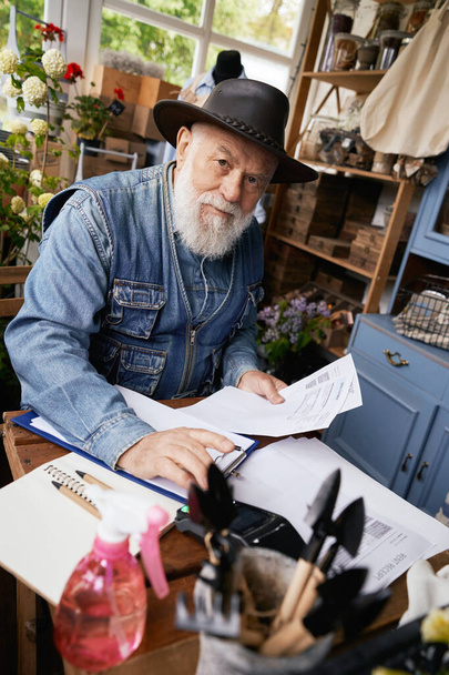 Vista superior de la cintura hacia arriba retrato de hombre de edad avanzada en sombrero sentado en el escritorio y trabajando con papeles y terminal de punto de venta - Foto, imagen