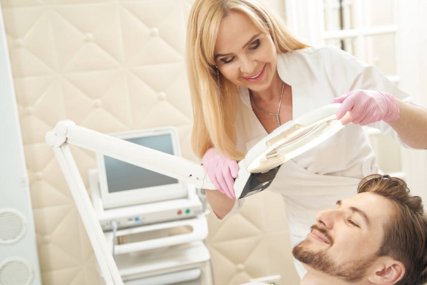 Smiling beautician in aesthetic clinic moving surgical light and illuninating face of man during beauty procedure - Photo, Image
