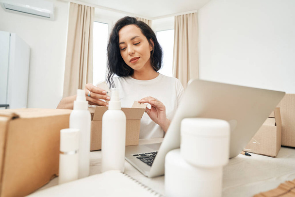Focused woman sitting at table and putting beauty product into open cardboard box - Foto, immagini