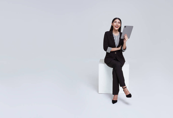 przyjazny twarz Azji bizneswoman uśmiech w formalny garnitur siedzi na krześle jej za pomocą tabletu i patrząc na copyspace izolowane na białym tle studio strzał. - Zdjęcie, obraz