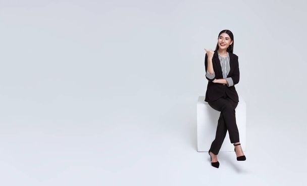friendly face asian businesswoman smile in formal suit sitting on chair and points her hands presented to copy space on white background studio shot. - Fotó, kép