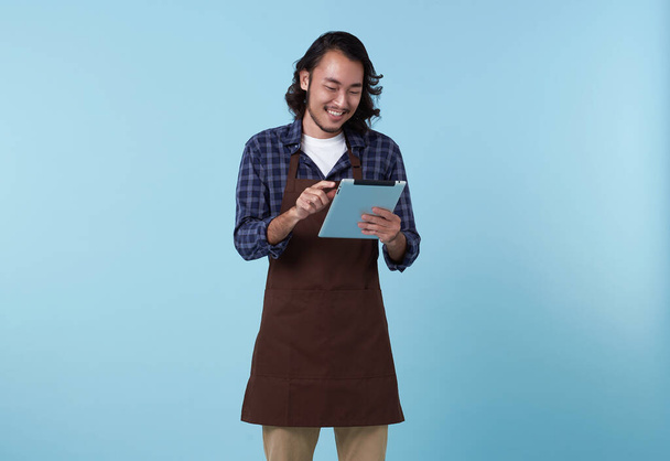 Jonge Aziatische ondernemer in schort hand holding tablet voor lijst bestellen voedsel geïsoleerd op blauwe achtergrond. Barista en serveerster concept. - Foto, afbeelding