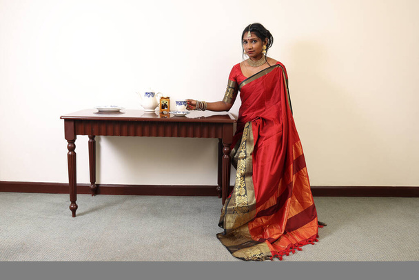 Indická žena na sobě červené oranžové tradiční královské saree a šperky umístění vinobraní stůl čaj hrnec šálek jasné okno podsvícení - Fotografie, Obrázek