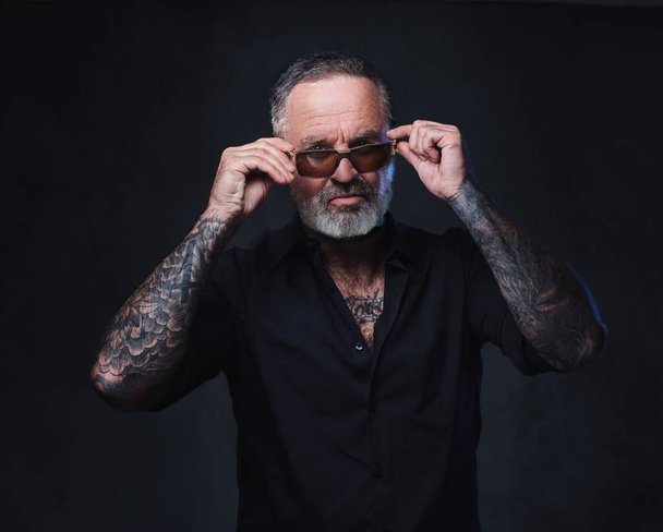 Studio shot of brutal aged man with sunglasses and tattooed body against dark background. - Fotoğraf, Görsel