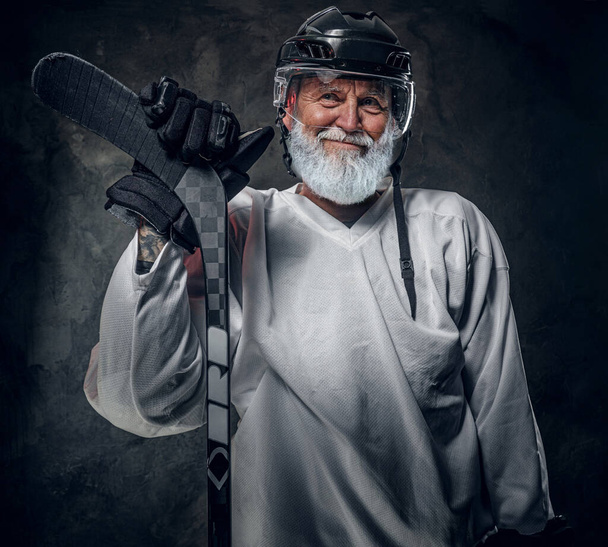 Photo d'un homme âgé joyeux vêtu de vêtements blancs sportifs et d'un casque tenant un bâton de hockey. - Photo, image