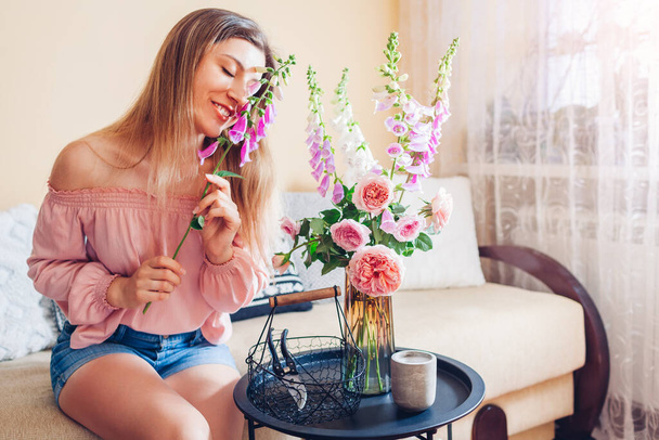 Woman smells flowers arranging bouquet with roses and foxgloves at home. Fresh blooms put in vase on table. Interior and summer decor - Photo, Image