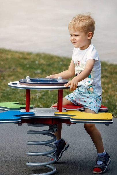 De vrolijke jongen springt op en balanceert op de schommels van een moderne buitenspeeltuin. - Foto, afbeelding
