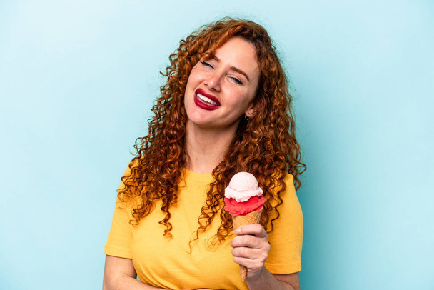 Young ginger woman holding an ice cream isolated on blue background laughing and having fun. - Photo, Image