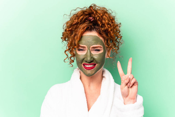 Mujer de pelo rojo joven con una máscara facial de aloe vera después de un baño aislado mostrando el número dos con los dedos. - Foto, Imagen