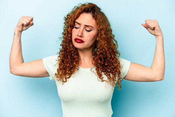 Young ginger caucasian woman isolated on blue background showing strength gesture with arms, symbol of feminine power - Photo, Image