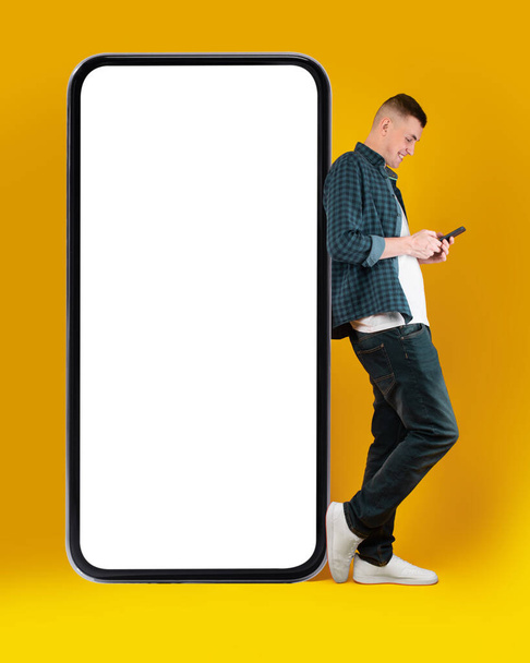 Millennial Man Using Phone Standing Leaning On Huge Smartphone Posing Over Yellow Studio Background. Great Mobile Application Advertisement Concept. Vertical Shot, Mockup - Photo, Image