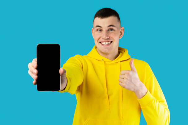 Great App. Man Showing Phone And Thumbs Up Gesture Approving Mobile Offer Smiling Looking At Camera Posing Over Blue Studio Background. I Like This Application For Cellphone. Mockup - Photo, Image