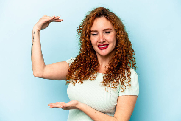 Young ginger caucasian woman isolated on blue background holding something little with forefingers, smiling and confident. - Foto, afbeelding