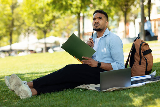 Concepto de educación online. Estudiante afroamericano reflexivo que estudia al aire libre, usando el ordenador portátil y tomando notas, sentado en el parque de la ciudad. Ocio estudiantil y estilo de vida. - Foto, imagen
