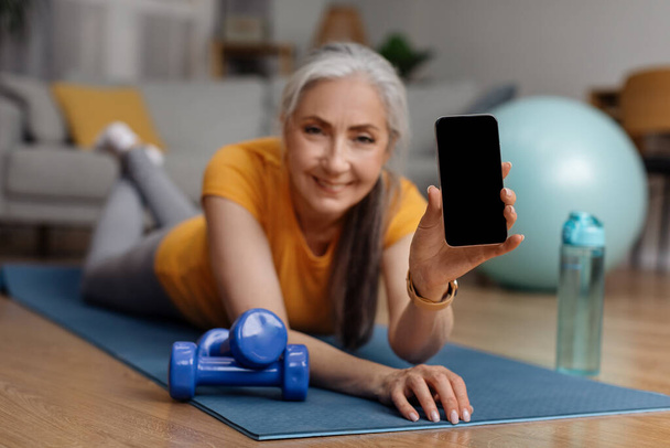 Mujer mayor feliz acostada en una esterilla de yoga y mostrando un teléfono inteligente con pantalla en blanco, utilizando la aplicación móvil de fitness mientras hace ejercicio en casa, maqueta, espacio para copiar - Foto, imagen