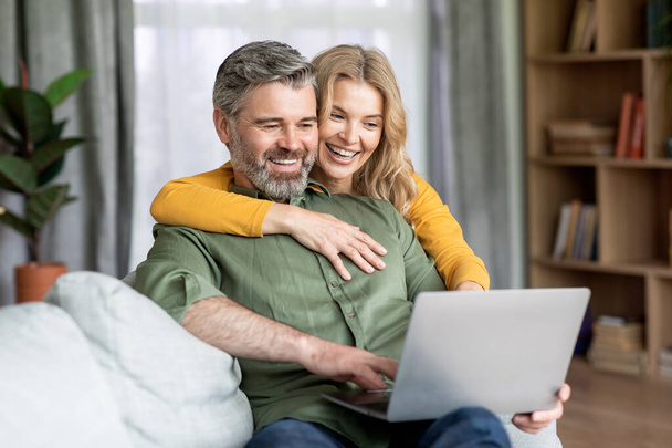Modern Technologies. Joyful Middle Aged Spouses Spending Time With Laptop At Home, Smiling Mature Couple Relaxing In Living Room With Computer, Browsing Internet Or Shopping Online, Free Space - 写真・画像