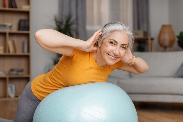 Mujer mayor feliz haciendo ejercicios con pelota de fitness en casa, entrenando sus músculos de la espalda con equipo deportivo durante el entrenamiento doméstico, señora mirando y sonriendo a la cámara - Foto, Imagen