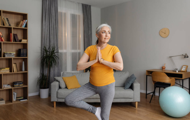 Domestic yoga practice. Senior woman standing in tree pose, keeping balanced, training at home in living room. Happy female in sportswear exercising indoors, copy space - Photo, Image