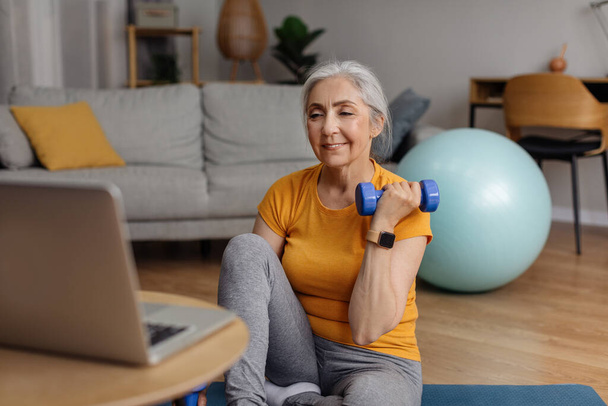 Domestic online sports training. Senior woman doing exercises with dumbbells in front of laptop in living room. Fit mature lady working on her arm muscles, following video tutorial - Fotó, kép
