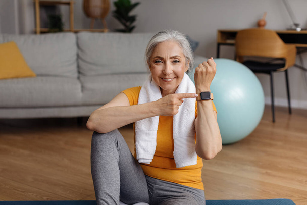 Happy fit senior woman pointing at activity fitness tracker and smiling at camera, having break in her training at home. Modern sports gadgets, fitness equipment concept - Photo, Image