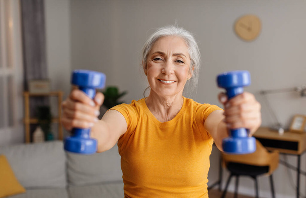 Home sports. Happy senior woman doing fitness exercises with weights, holding dumbbells and training at home. Aged lady strengthening her arm muscles, leading healthy lifestyle - Photo, Image
