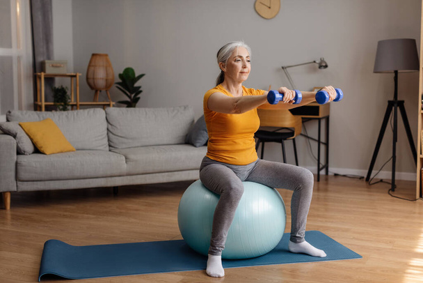 Active senior woman sitting on fitness ball and working out with dumbbells, exercising in living room at home, copy space. Aged lady taking care of her health - Photo, Image
