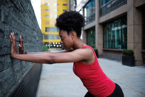 陸上競技の若い黒人女性がロンドンの壁に向かって伸びている。彼女は赤い袖のないTシャツを着ている。髪の毛. - 写真・画像
