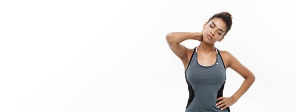 Υγιείς και γυμναστήριο έννοια - πορτρέτο της αφρικανικής αμερικανικής κορίτσι υποφέρει ένα μυϊκό τραυματισμό στέκεται κρατώντας το λαιμό και κάτω μέρος της πλάτης. Απομονωμένα σε λευκό φόντο - Φωτογραφία, εικόνα