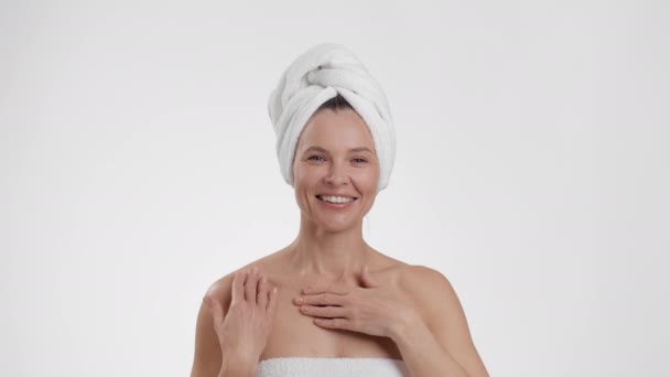 Μετά τις διαδικασίες spa ντους. Ευτυχισμένη μεσήλικη κυρία με πετσέτα στο κεφάλι απλώνοντας κρέμα περιποίησης στο σώμα της, ενυδατικό δέρμα μετά το μπάνιο, λευκό φόντο στούντιο, αργή κίνηση - Πλάνα, βίντεο