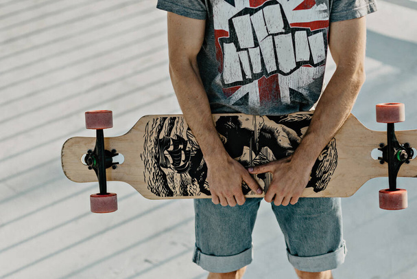 Τα αρσενικά χέρια έχουν ξύλινο σκέιτμπορντ. Κοντινό πλάνο ενός νεαρού που κρατάει ένα σκέιτμπορντ στα χέρια του, κάνοντας πατινάζ στο φόντο της φύσης. Δωρεάν χαλαρωτικό skateboard μοντέρνα έννοια. στυλ hipster - Φωτογραφία, εικόνα