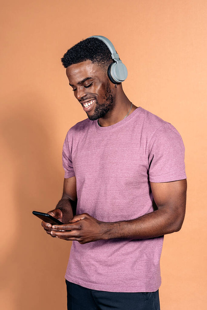 Archivfoto eines gut aussehenden schwarzen Mannes mit Kopfhörern, der im Studio vor braunem Hintergrund auf sein Handy schaut. - Foto, Bild