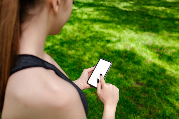 Piękna brunetka kobieta nosi sportowy biustonosz stojący w parku miejskim, na zewnątrz ręce trzymając telefon dotykając palec makieta biały pusty wyświetlacz, aplikacja mobilna tech koncepcja, przez ramię widok z bliska. - Zdjęcie, obraz