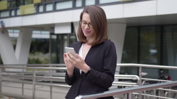 Content business woman in stylish black dress standing on a ganek of office building and reading message on smartphone during break - Materiał filmowy, wideo
