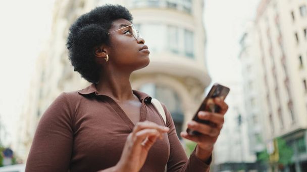 Dark-skinned Afro haired girl standing outdoors. Young thoughtful woman looking for right destination using navigation on smartphone - Photo, Image