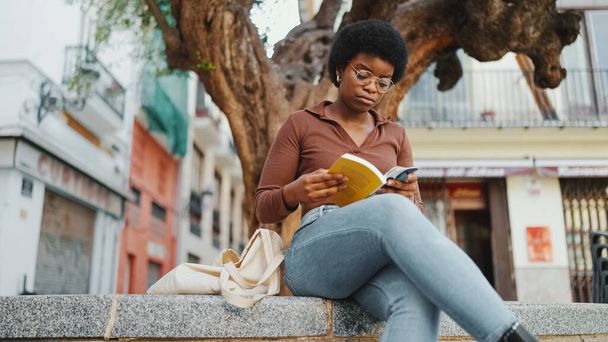 Young African woman looking concentrated while reading a book under the tree, wearing glasses. Attractive student girl studying outdoors - Photo, Image