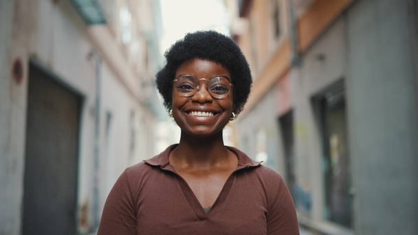 Iloinen afrikkalainen nainen silmälaseissa näyttää onnelliselta ulkona. Huoleton tummaihoinen tyttö hymyilee vilpittömästi kadulla. Tyttö katselee kameraa - Valokuva, kuva