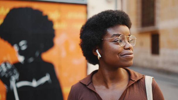 Side view of attractive Afro American woman in glasses looking happy exploring a new city with music in earphones - Photo, Image