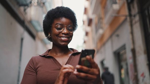 Молодая афро-девушка в очках выглядит счастливой, переписываясь с друзьями на смартфоне. Женщины используют мобильный телефон на открытом воздухе - Фото, изображение
