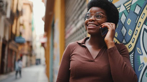 スマートフォンの屋外で話して魅力的なアフリカの女性。陽気な女の子を持っています会話上の友人と携帯電話 - 写真・画像